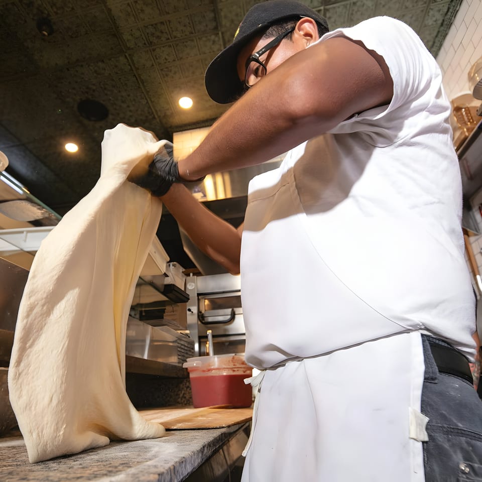 A pizza chef in a black cap and white apron stretches fresh pizza dough at Brooklyn DOP.