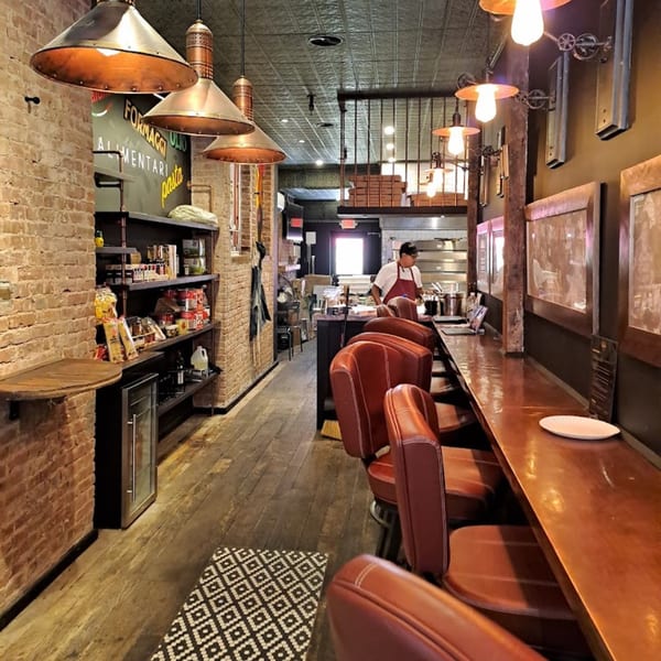 Interior of Brooklyn DOP, a pizza restaurant, with a long bar, stools, brick walls, and a chef in the background.