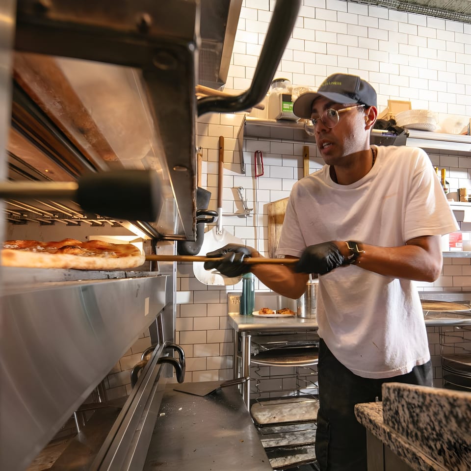 A Brooklyn DOP chef in a cap and glasses uses a peel to remove a pizza from a hot oven.