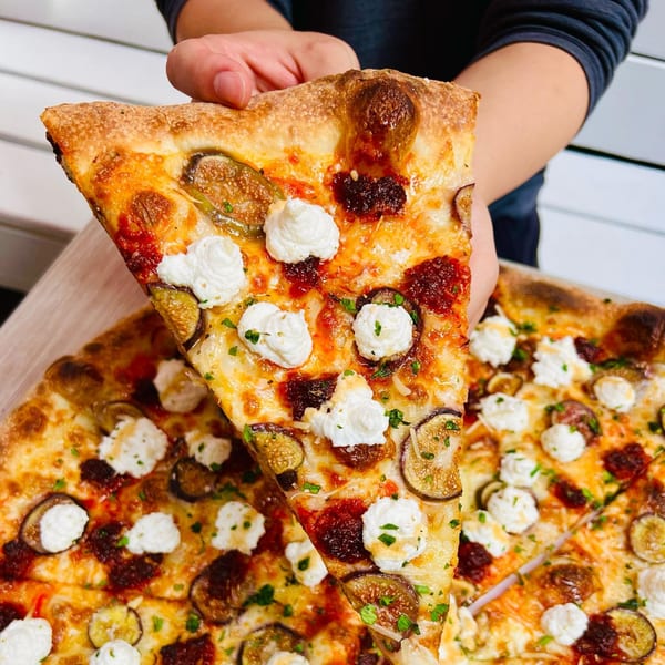 Hand holding a slice of Brooklyn DOP pizza with figs, ricotta, and red sauce over a full pizza.