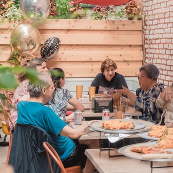 People enjoying pizza and drinks at an outdoor table with balloons and brick wall.