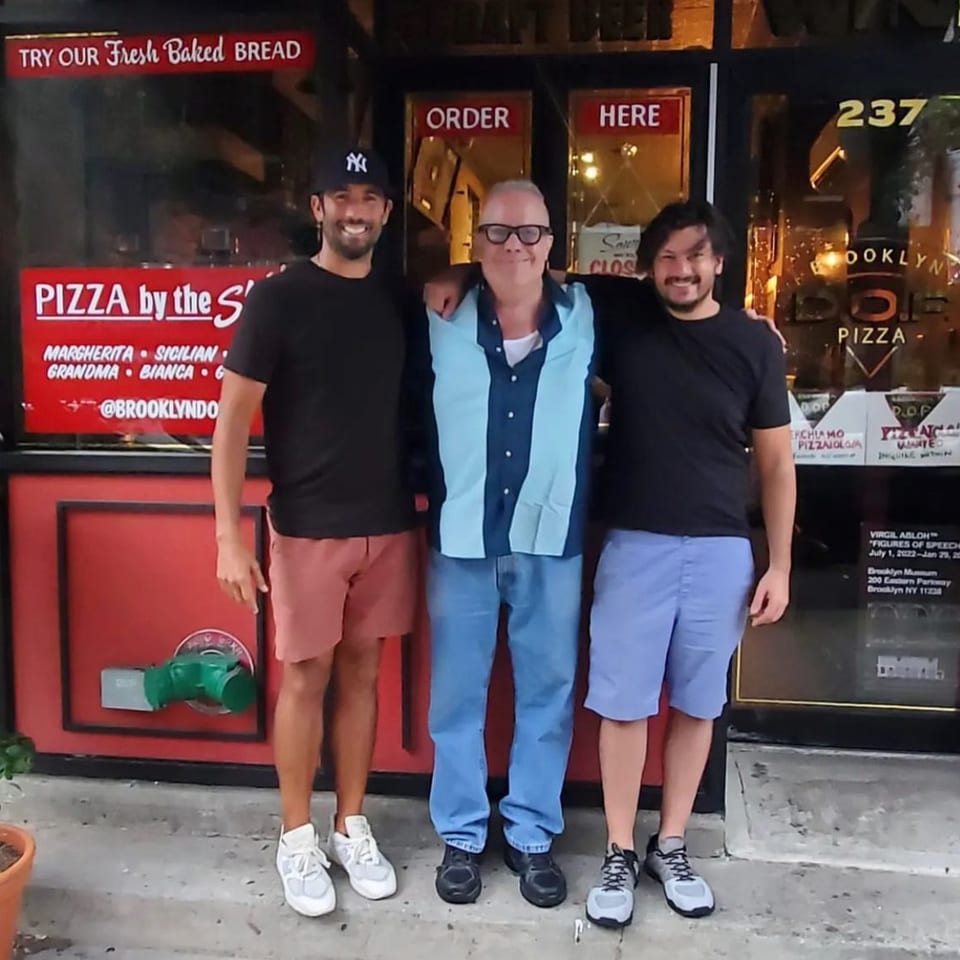 Three men smiling, standing in front of Brooklyn DOP pizza shop entrance.