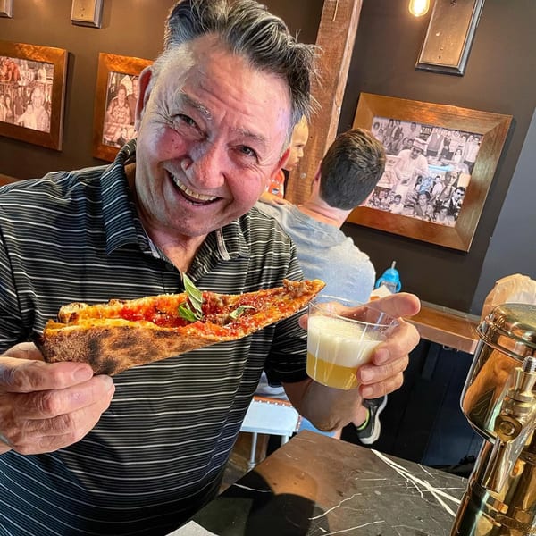 A smiling man holds a slice of Brooklyn DOP pizza and a drink in a lively restaurant.